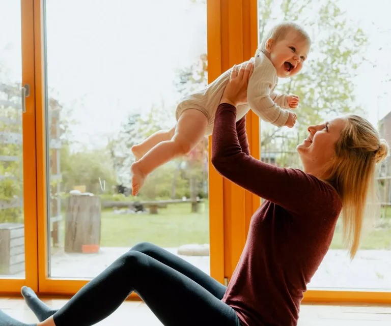 Eine junge Mutter hält sitzend ihr Baby hoch und beide lächeln.