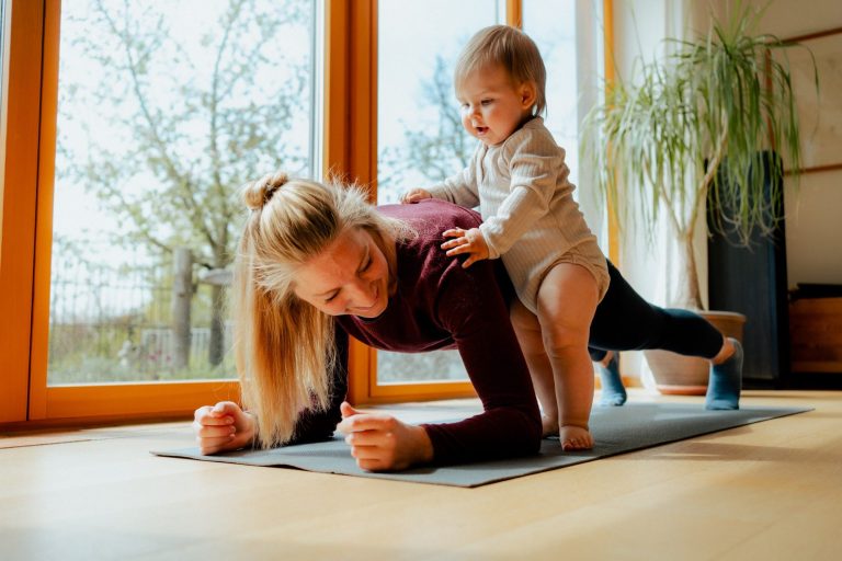 Frau führt die Sportübung Plank auf einer Matte vor einem Fenster aus und das Baby steht an ihrer Seite und hat die Hand auf ihrem Rücken